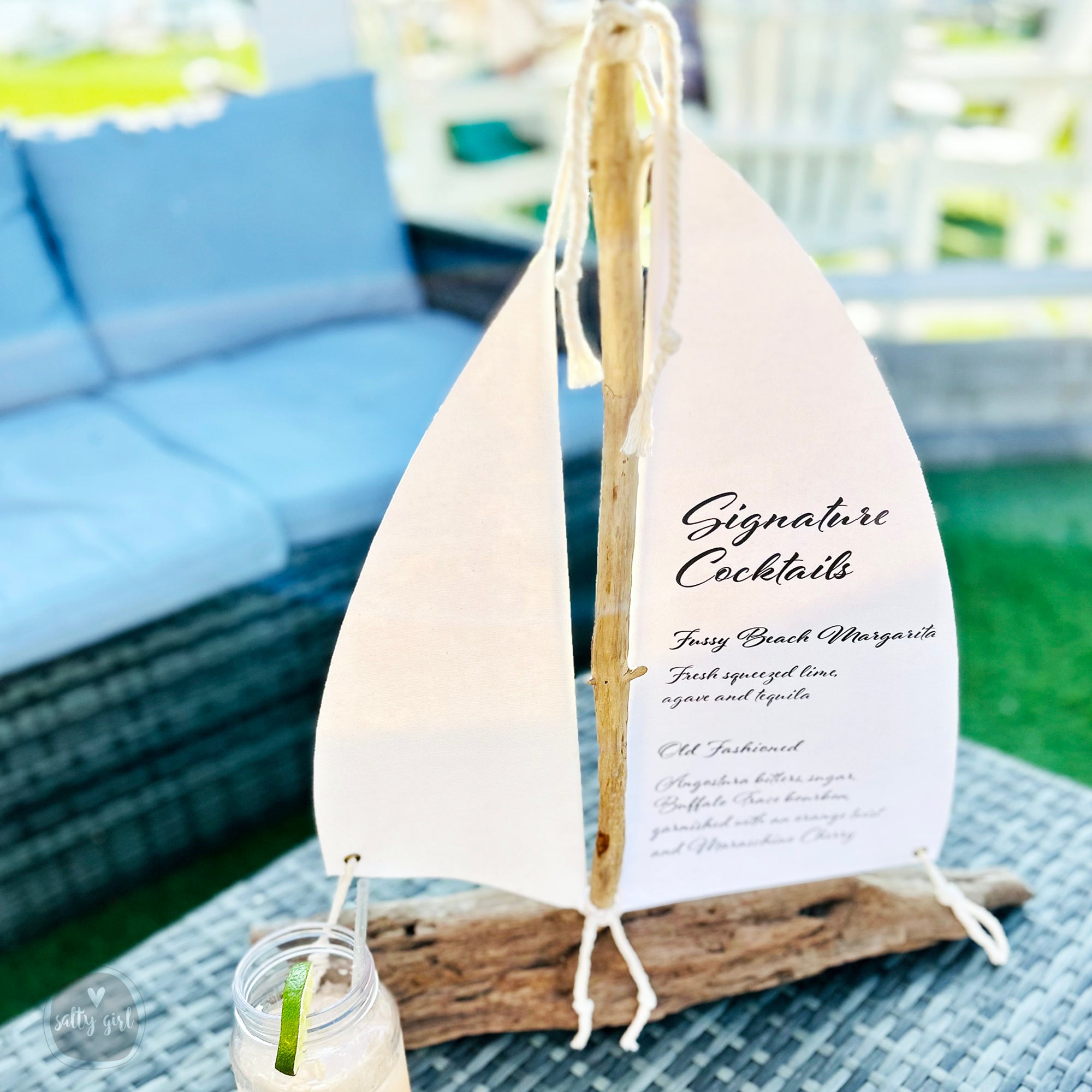 A white sailboat-shaped cocktail stand displaying a cocktail menu, placed on a table with a blue cushion and a glass of a tropical drink.
