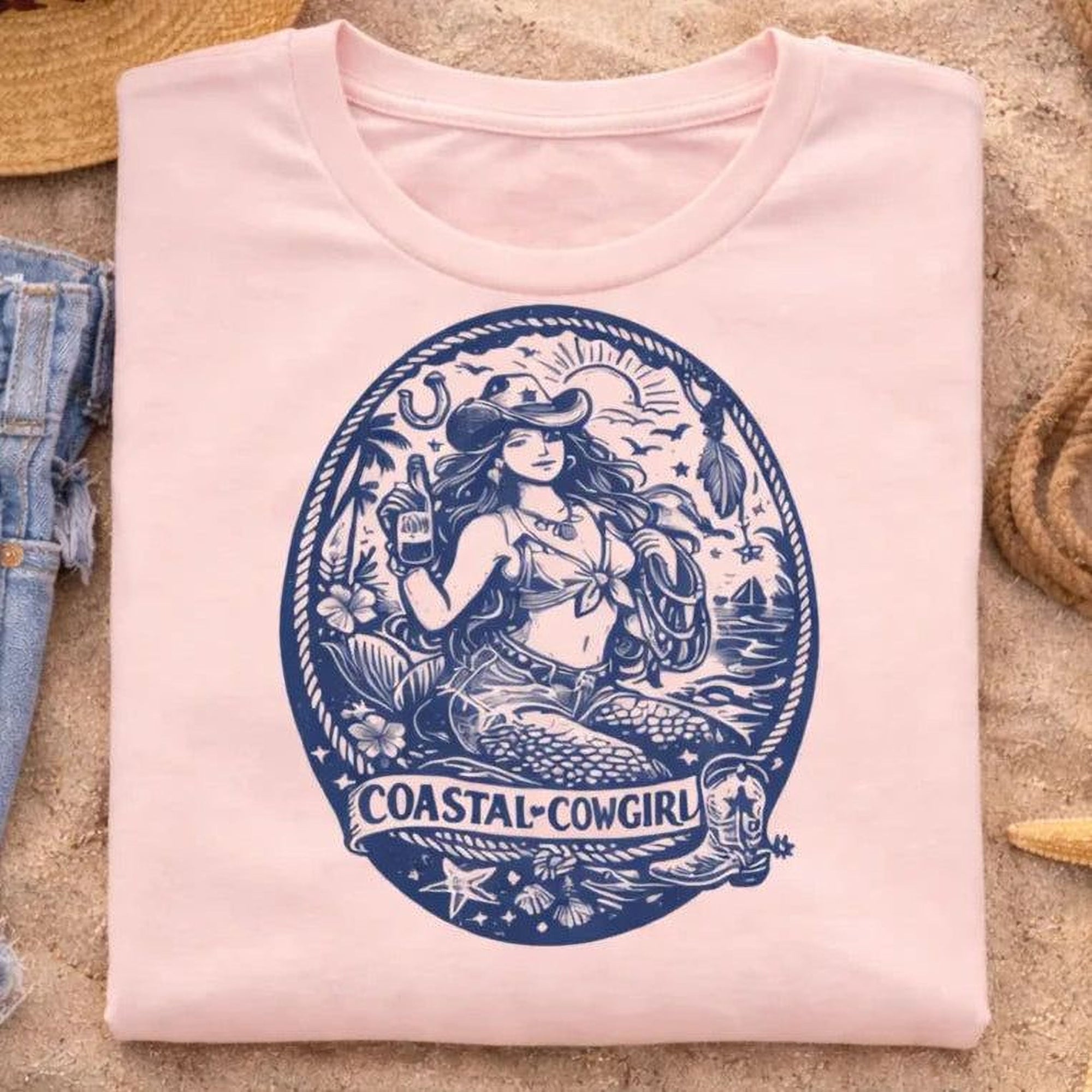A pink Maine Salty Girl t-shirt with a graphic design featuring a mermaid or mermaid-like figure, along with the text "COASTAL-COWGIRL" and various coastal elements such as a lighthouse, palm tree, and starfish.