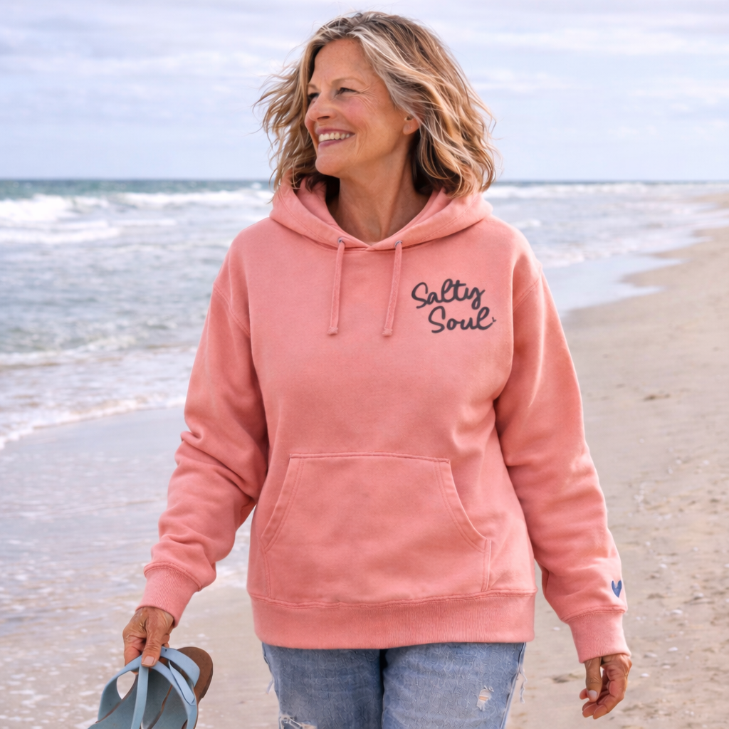 A woman wearing a pink hoodie with the text "Salty Soul" is walking on a beach, smiling and holding a flip-flop.
