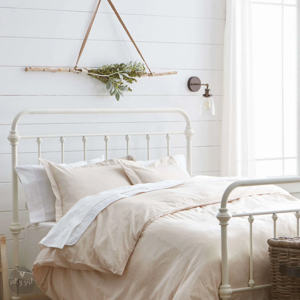 a cozy bedroom with a white metal bed frame, a white headboard, and a hanging plant arrangement. The room has white walls and a window, creating a bright and airy atmosphere.