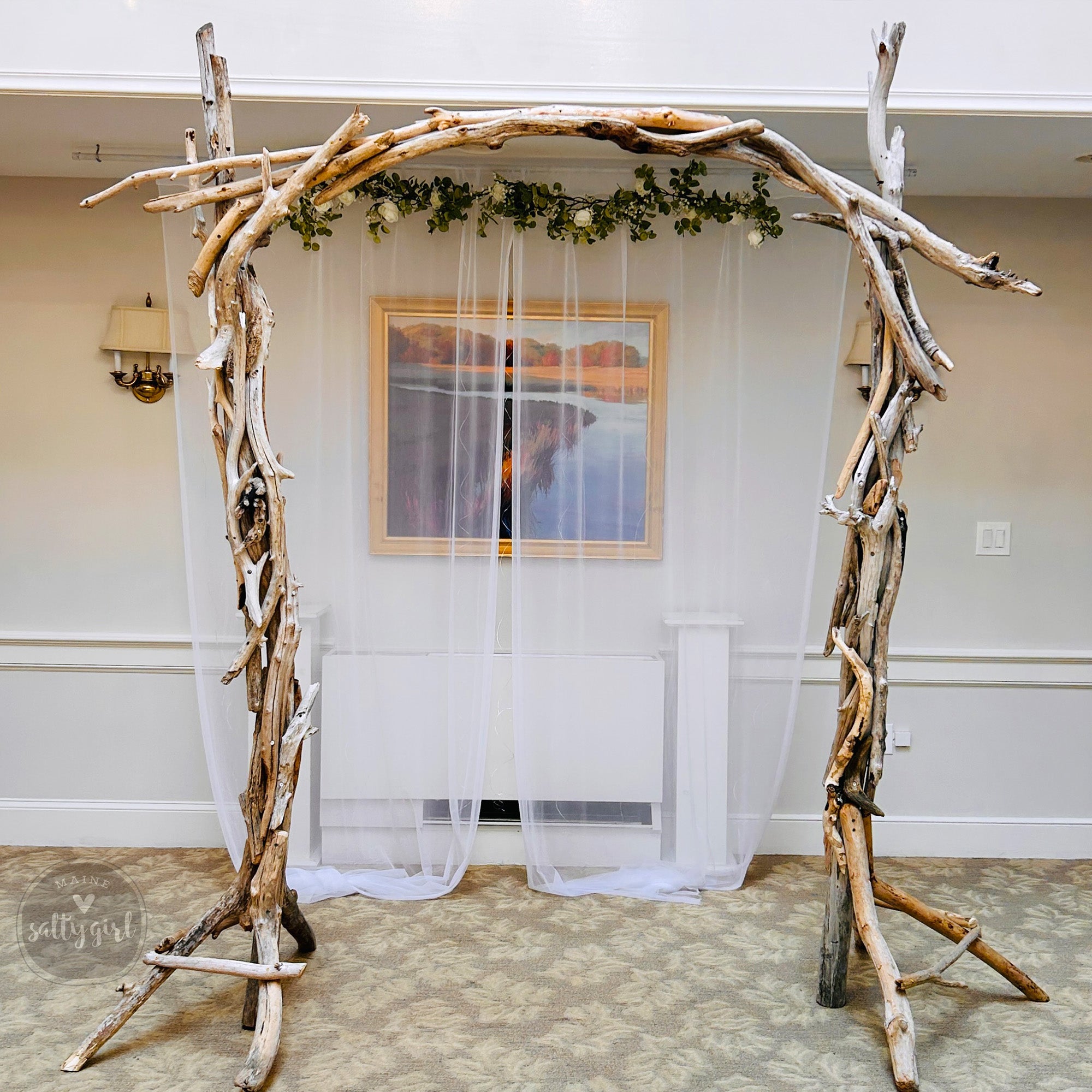 a rustic, natural-looking arch made of driftwood and branches, with a white curtain draped over it. The arch is situated in a room with a painting hanging on the wall behind it.