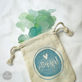 A beige cloth bag with a blue and white design, containing a pile of aqua sea glass.