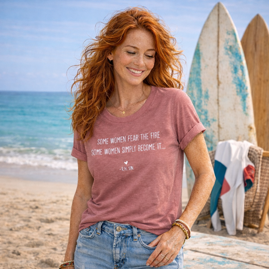 A woman with long red hair stands on a beach, smiling and wearing a pink t-shirt with a message about women facing the fire.