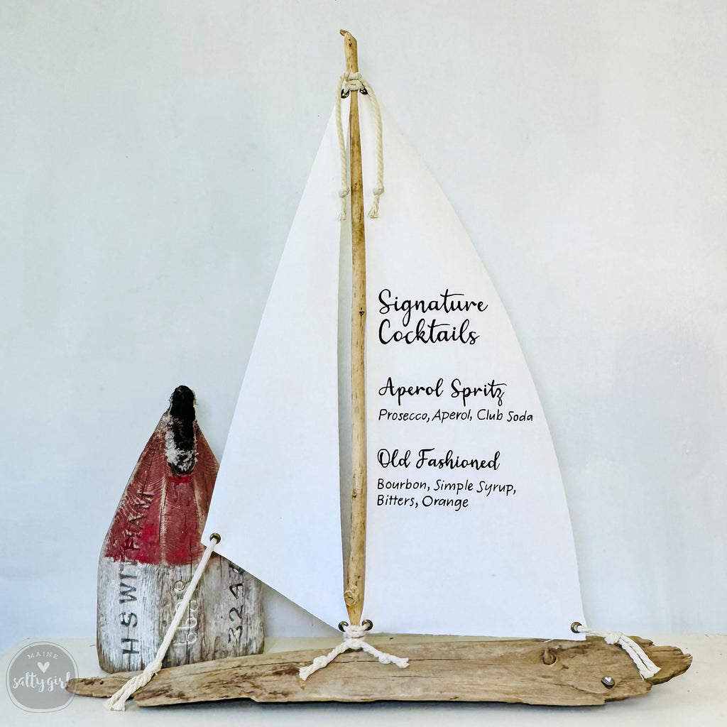 a wooden sailboat-shaped object with a white sail, displaying a list of signature cocktails.