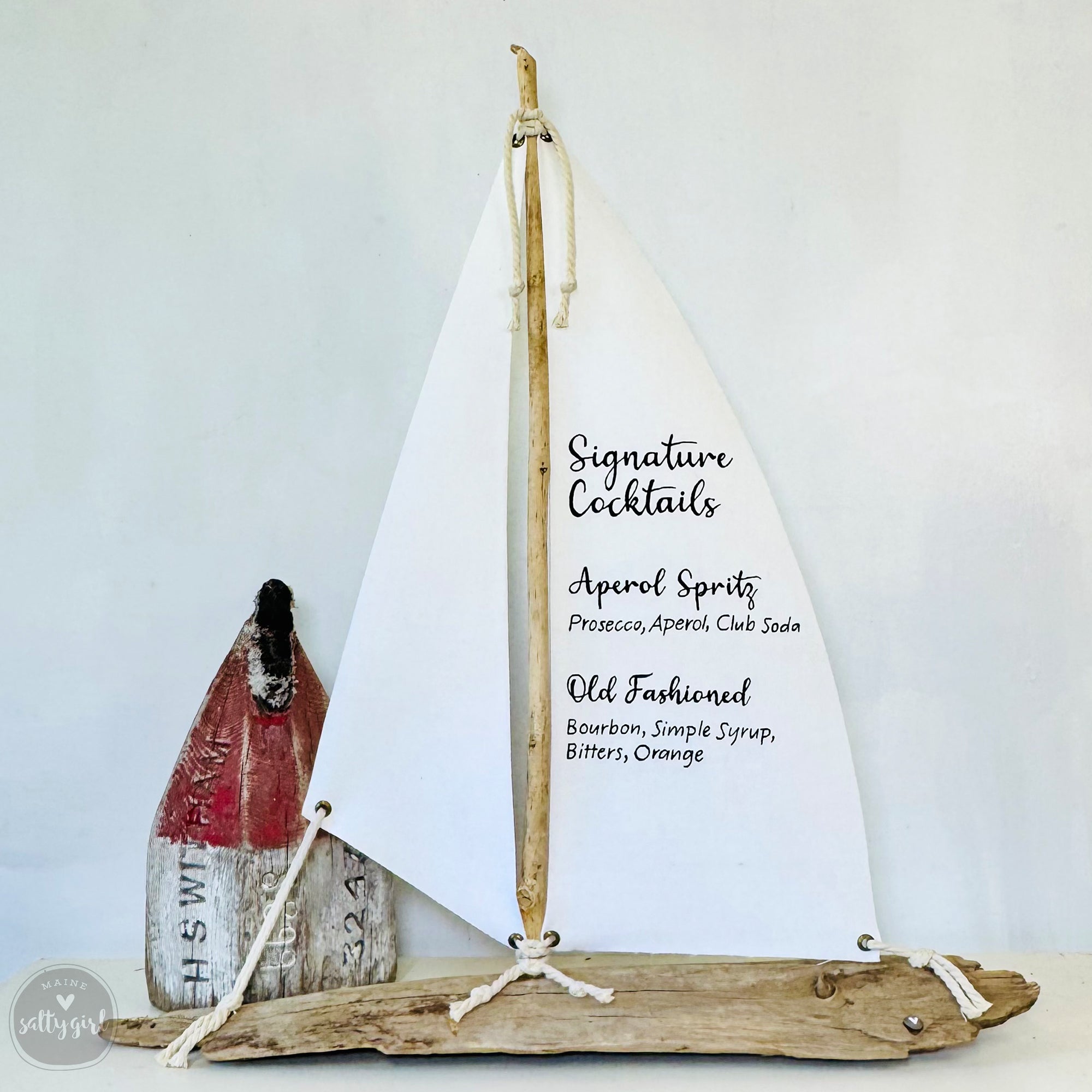 a wooden sailboat-shaped object with a white sail, displaying a list of signature cocktails.