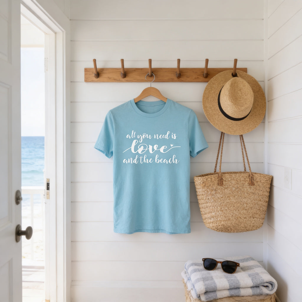 A blue t-shirt with the text "all you need is love and the beach" is hanging on a wooden coat rack, with a straw hat and a woven basket nearby.