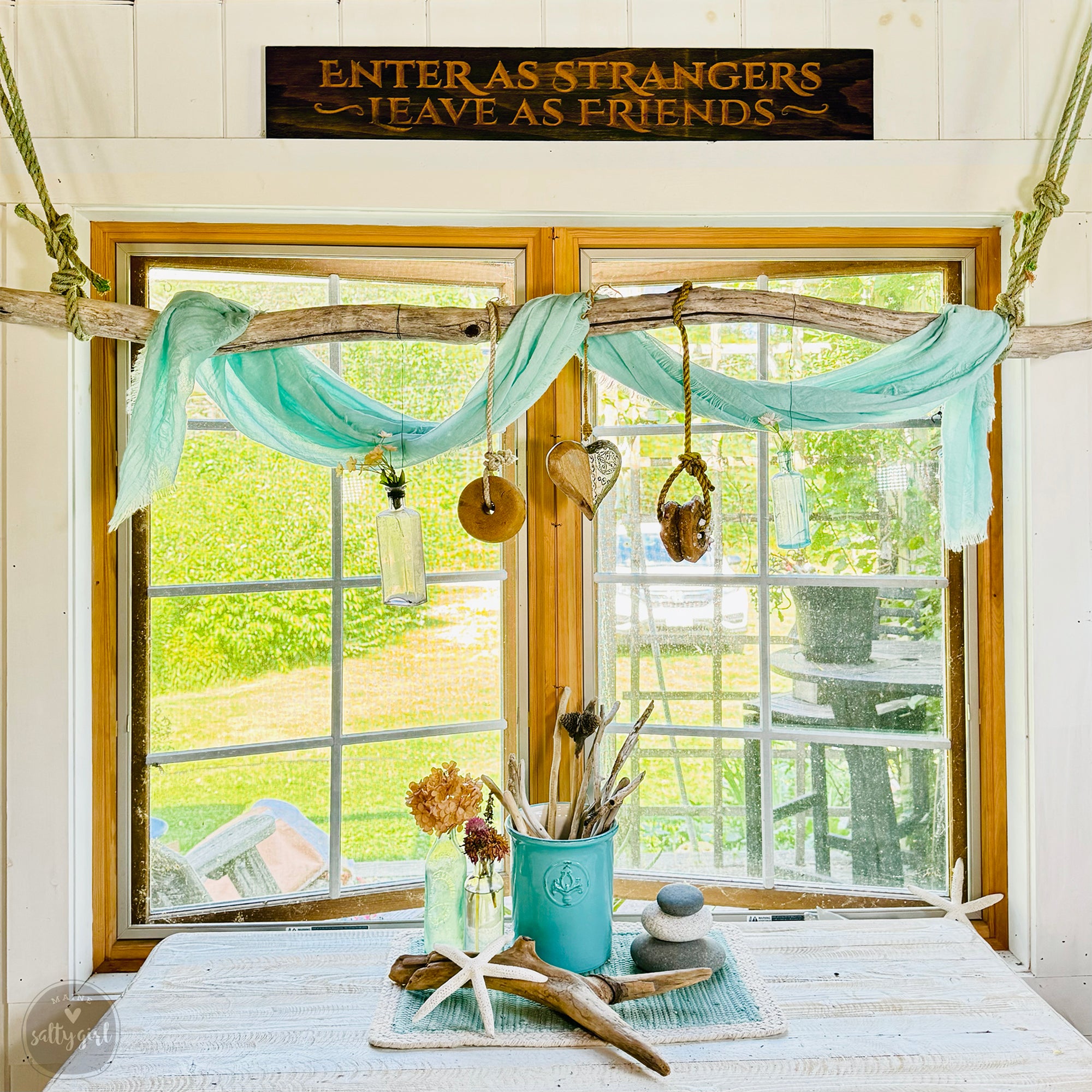 a window with a sign that reads "Enter as strangers, leave as friends", and a table with various objects on it, including a vase with flowers, a small sculpture, and a rock.