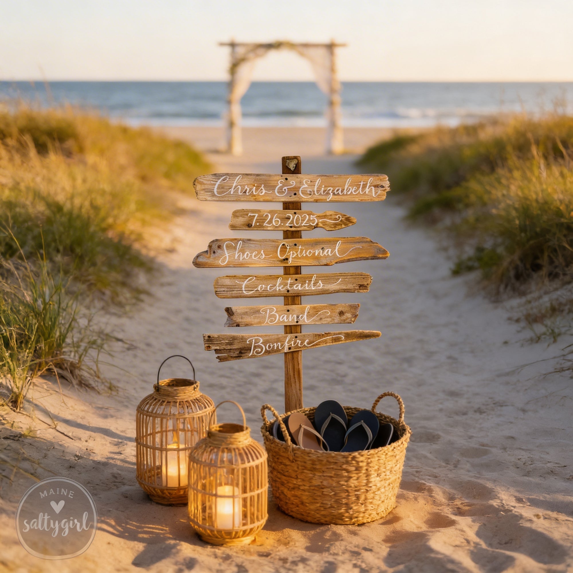 A rustic wooden signpost on a sandy beach, with two lanterns and a wicker basket filled with flip-flops, indicating a beach wedding or event.