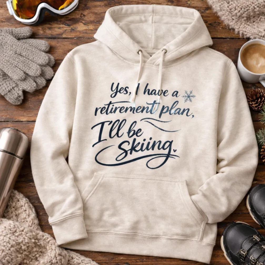 A beige hoodie with a humorous retirement plan and skiing message, along with ski gear and accessories, including gloves, a cup, and a pair of boots.