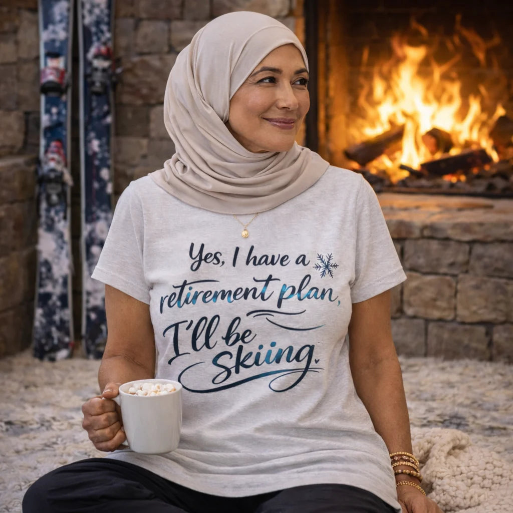 A woman wearing a hijab and a t-shirt with a humorous retirement plan is sitting in front of a fireplace, holding a mug of hot chocolate.