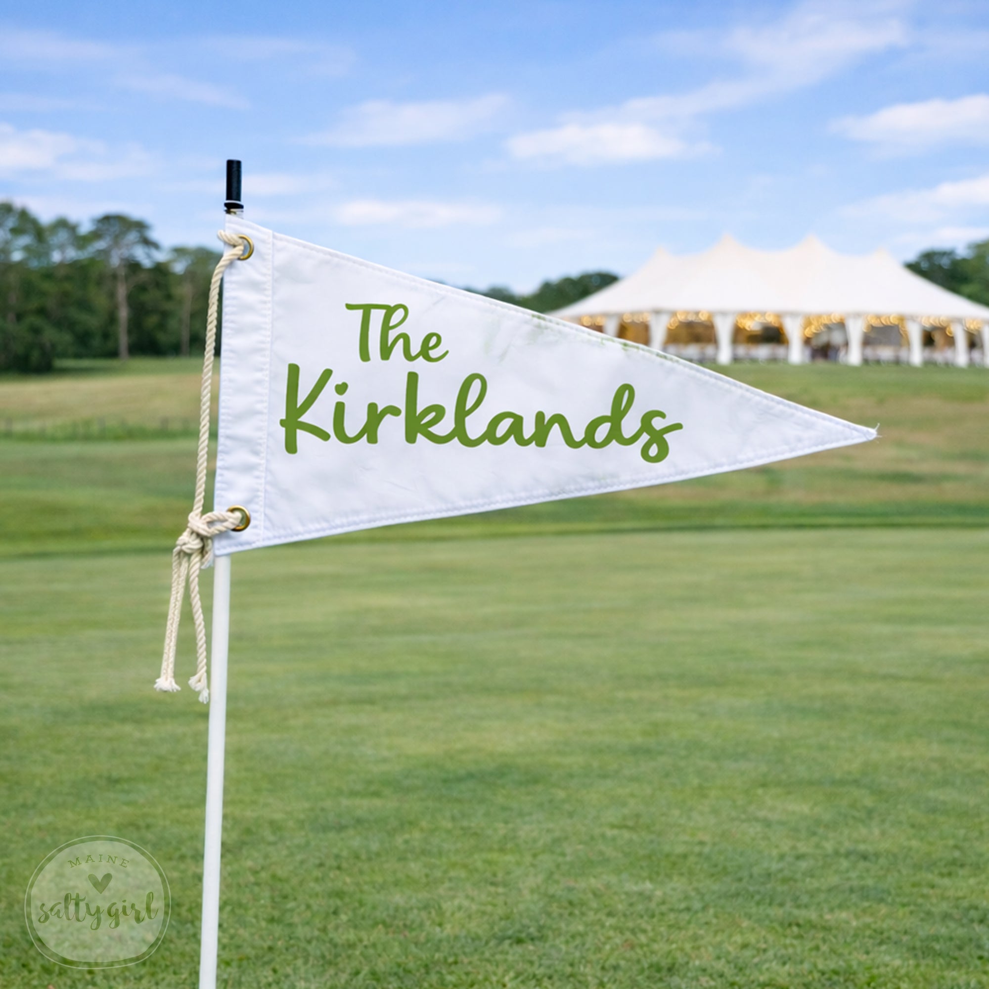 A white triangular flag with the text "The Kirklands" written on it, standing on a grassy field with a white tent visible in the background.