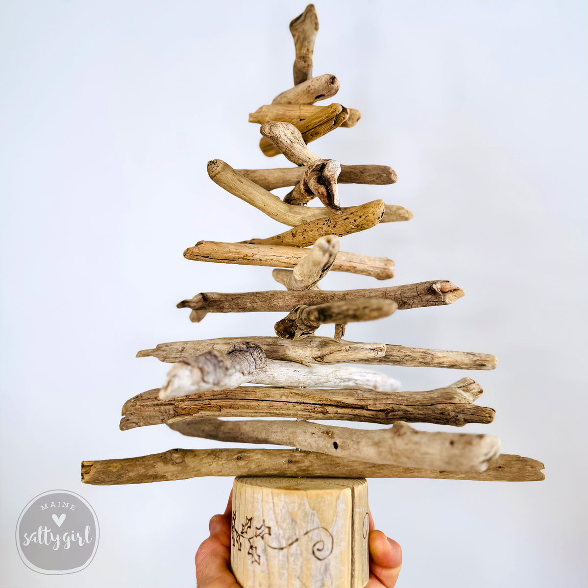 A hand holds a wooden base with a tree-like structure made of various wooden sticks and branches, creating a rustic and natural aesthetic.