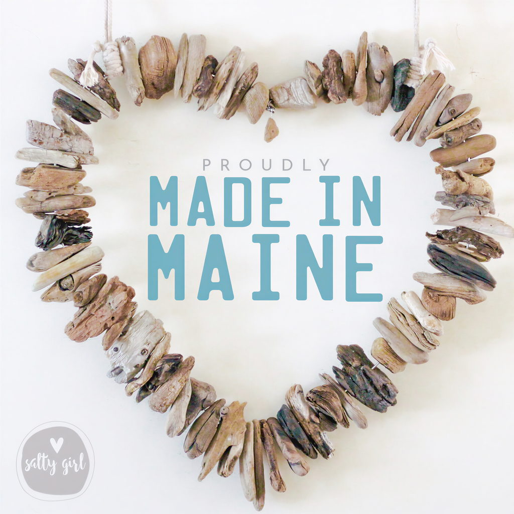 A heart-shaped arrangement of various natural materials, including small rocks, shells, and driftwood, with the text "PROUDLY MADE IN MAINE" prominently displayed in the center.
