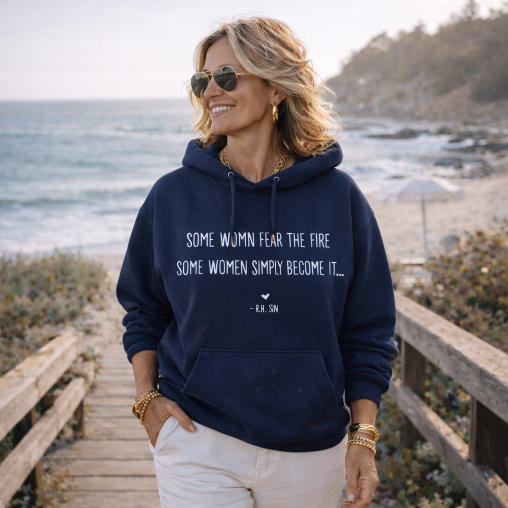 A woman wearing a navy blue hoodie with a quote printed on it, standing on a wooden boardwalk near the ocean.