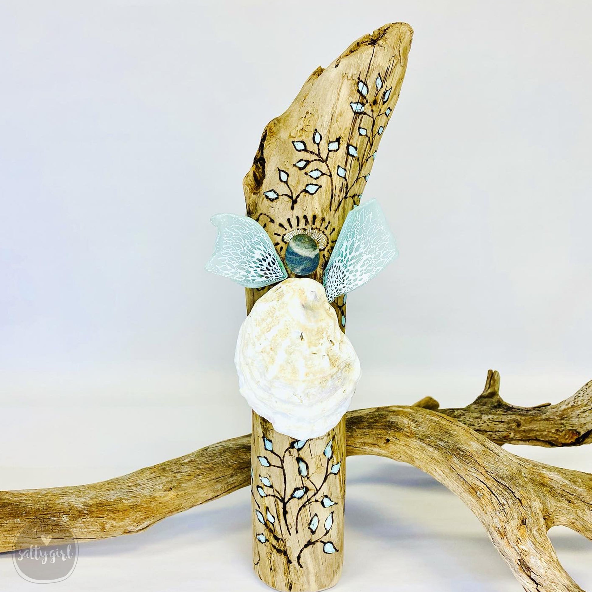 A handcrafted wooden angel figure made from driftwood, adorned with blue and white leaf patterns, and featuring a seashell at its base.