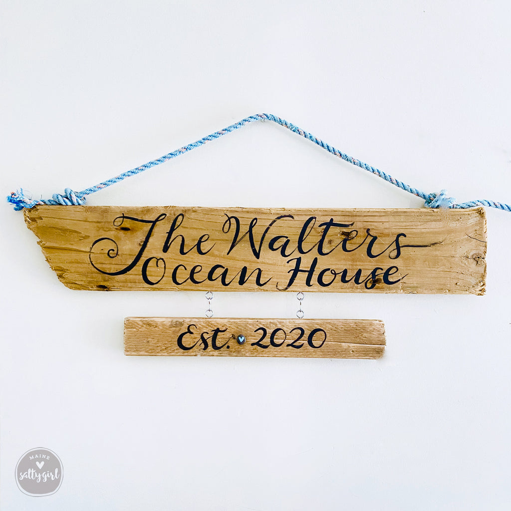 A wooden driftwood sign with the name "The Walters Ocean House" written on it, hanging on a rope.