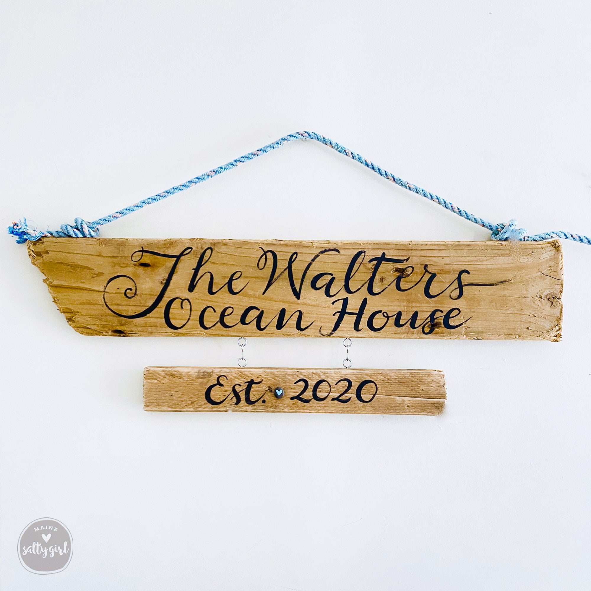 A wooden driftwood sign with the name "The Walters Ocean House" written on it, hanging on a rope.