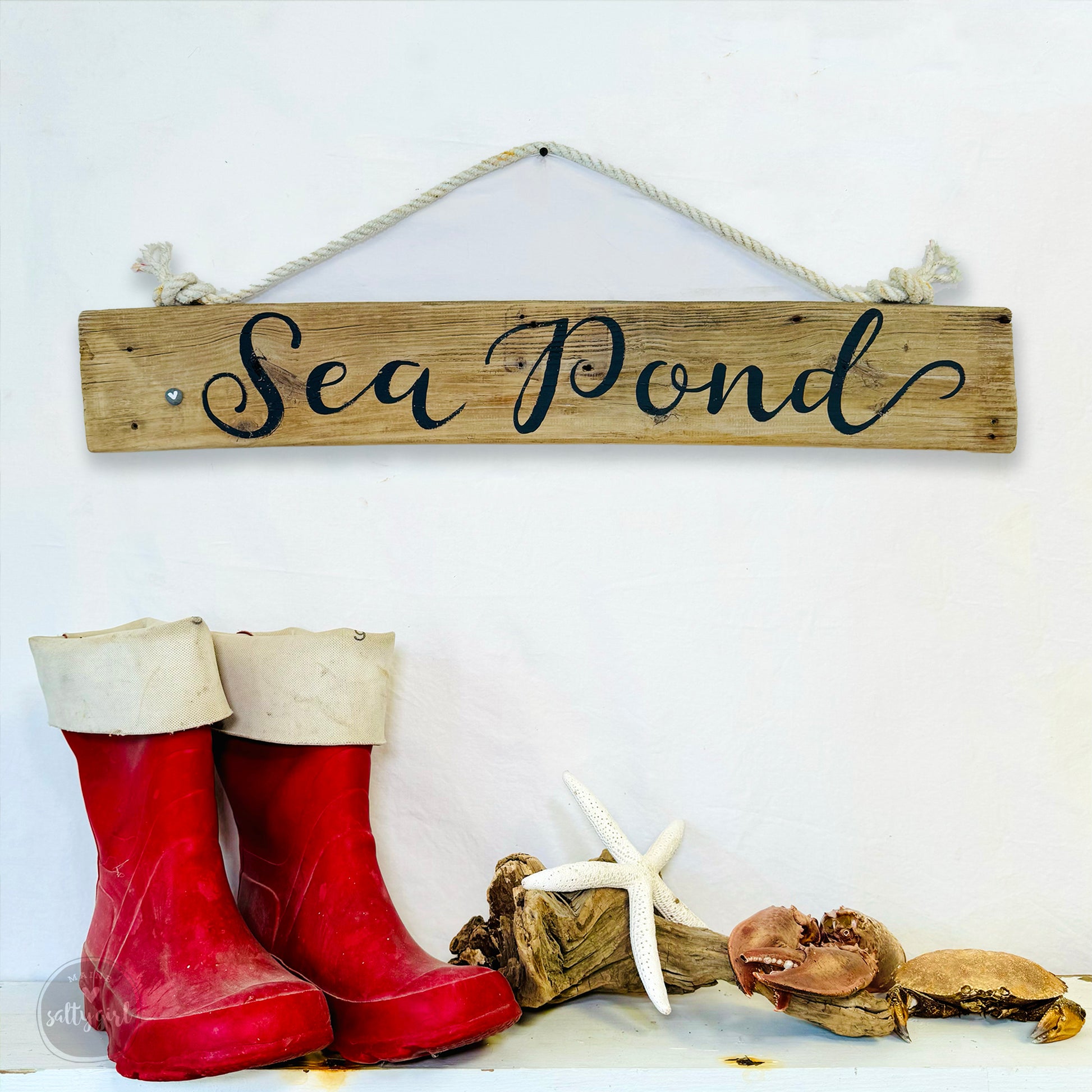 a rustic wooden sign with the word "Sea Pond" written on it, hanging on a white wall. Below the sign, there are two pairs of red rubber boots and a white starfish, creating a coastal-themed display.