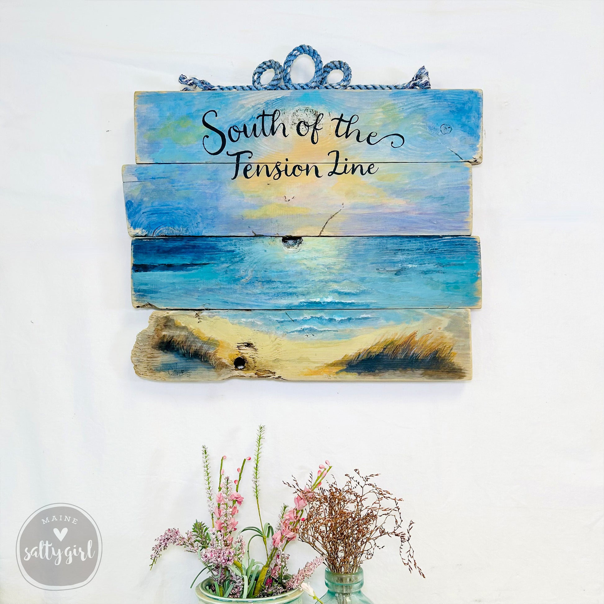 a wooden sign with a painting of a beach scene, hanging on a wall. Below the sign, there are two small vases with flowers, creating a decorative arrangement.