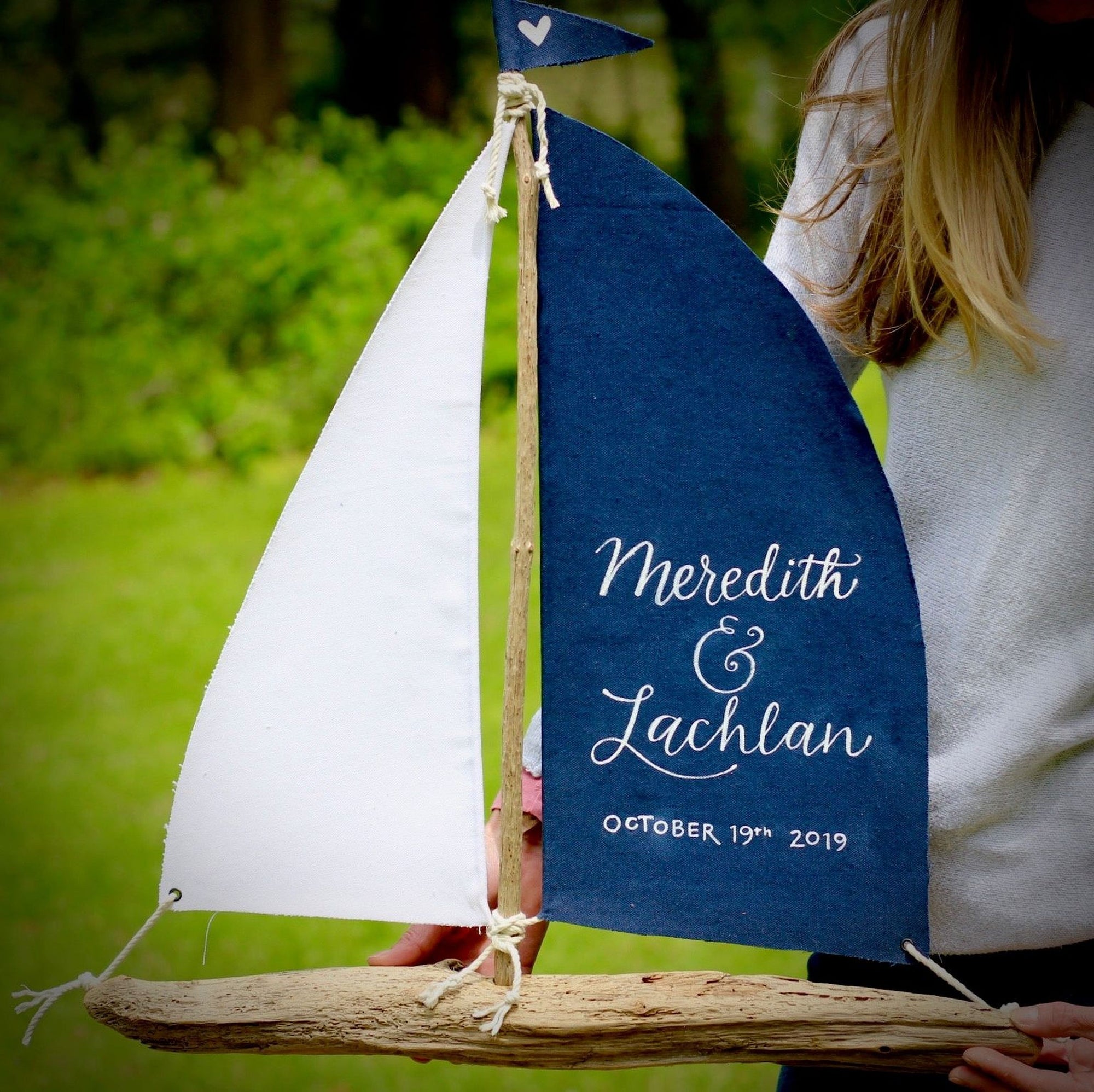 A person is holding a blue sailboat-shaped wedding invitation with white text, standing in a grassy area.