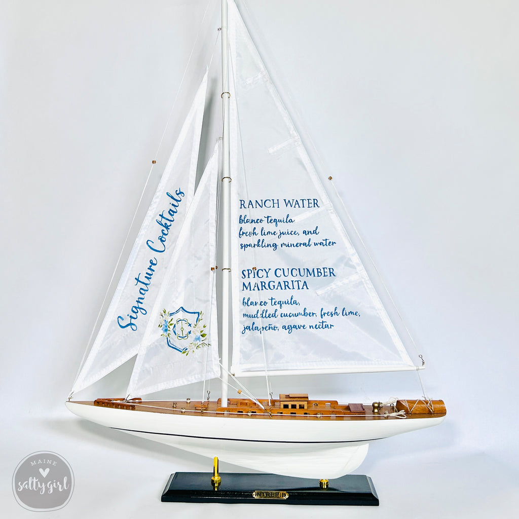 A model sailboat with a white hull and blue sails, displaying a list of signature cocktails on its sails.