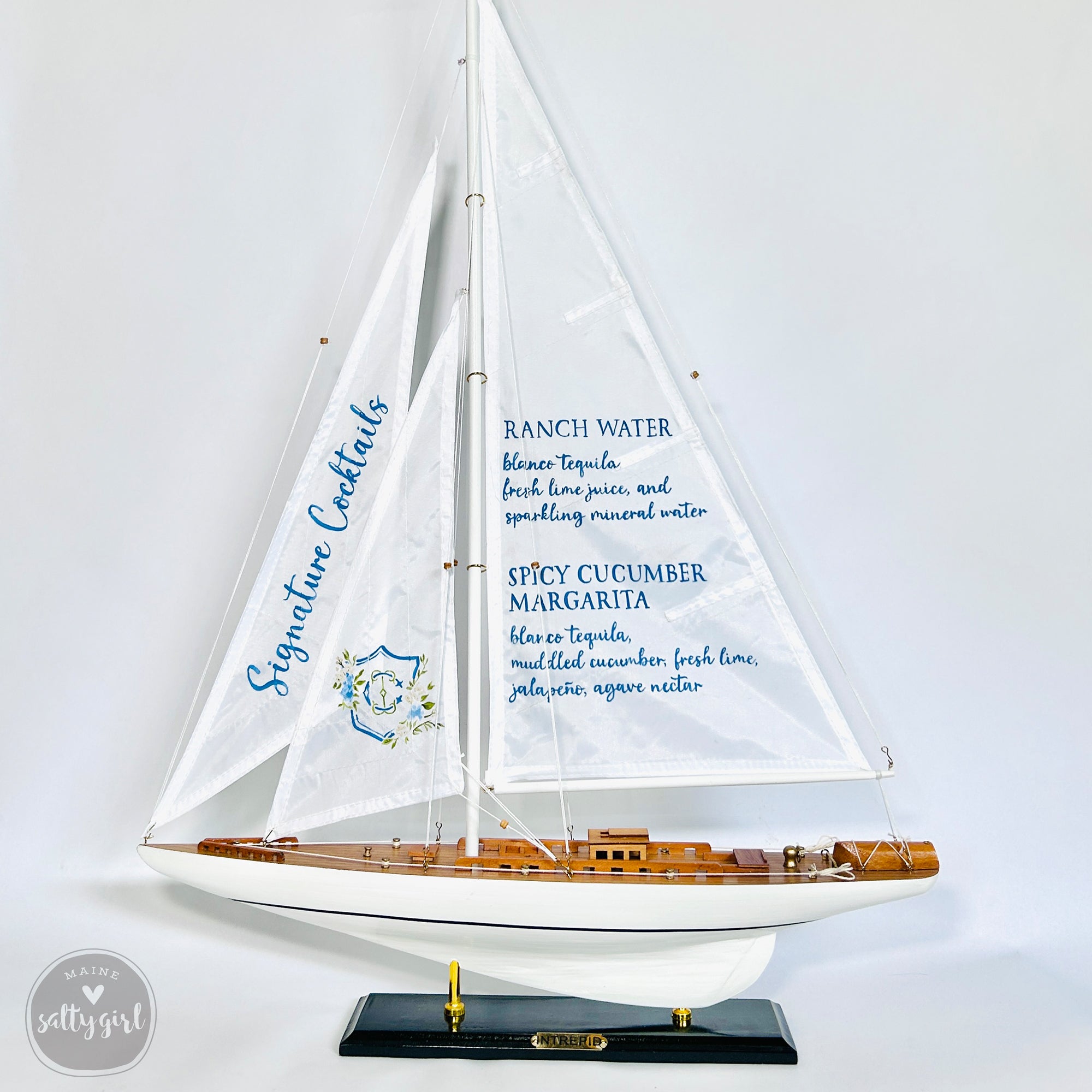 A model sailboat with a white hull and blue sails, displaying a list of signature cocktails on its sails.
