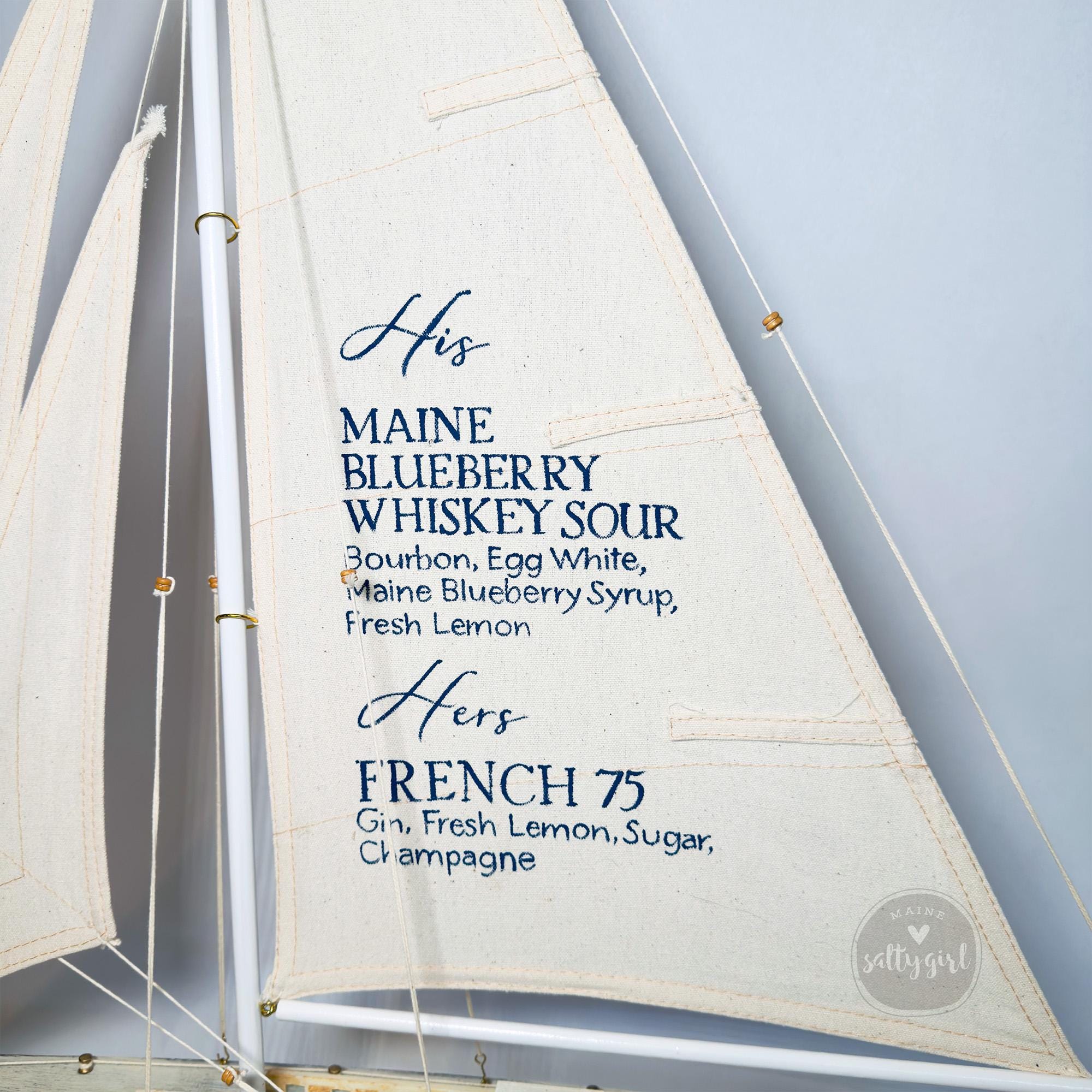 a close-up view of a sailboat's sail, with text printed on it. The text appears to be a menu or list of drinks, including "His" and "Her" along with their respective names, such as "MAINE BLUEBERRY WHISKEY SOUR", "BOURBON, Egg White", "MAINE BLUEBERRY SYRUP", "FRESH LEMON", and "FRENCH 75".