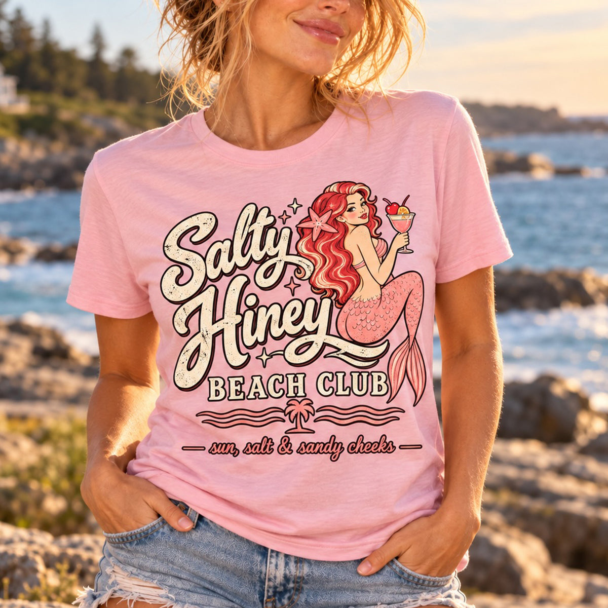 A woman wearing a pink t-shirt with a mermaid graphic and text that says "Salty Hiney Beach Club" and "sun, salt & candy cheeks".