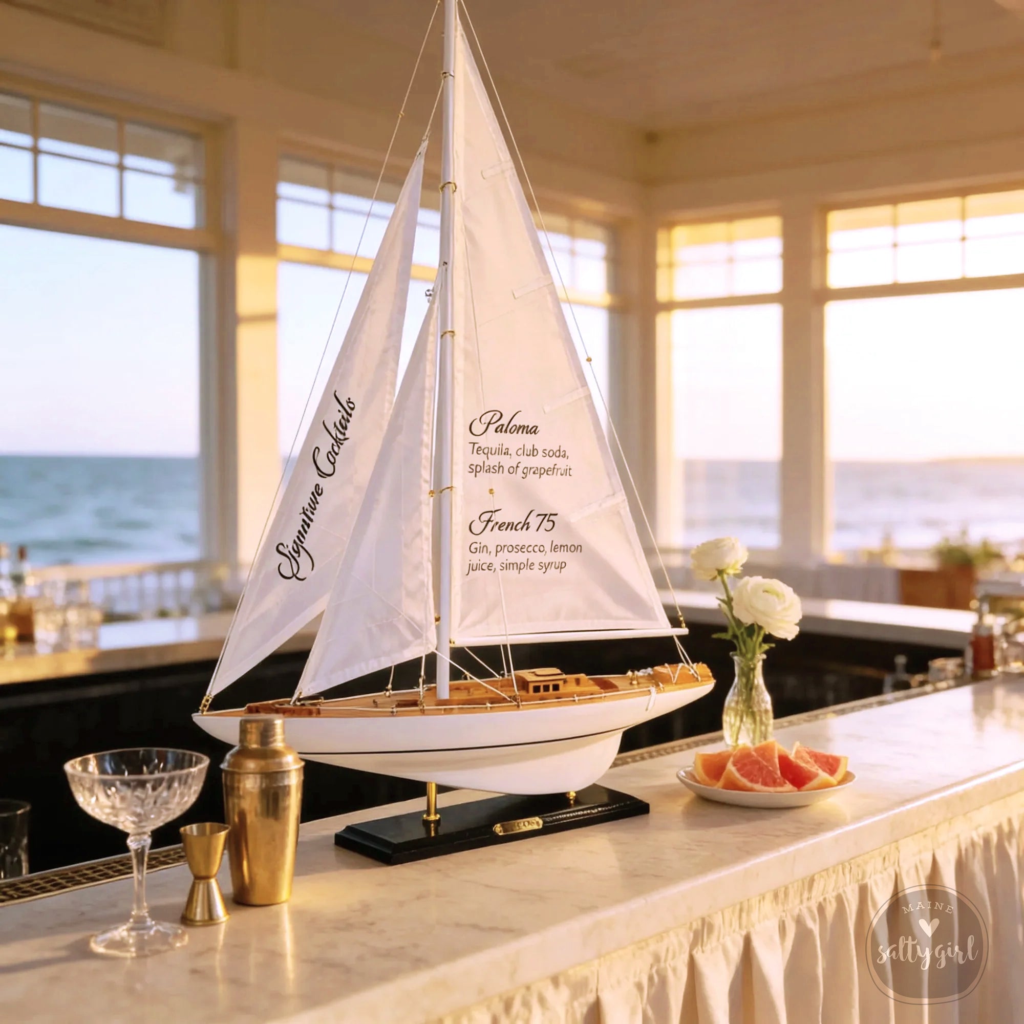a white Hand-Lettered Model Sailboat Bar Menu displayed on a bar counter, with a plate of fruit, a cocktail shaker, and a vase of flowers nearby. The sailboat