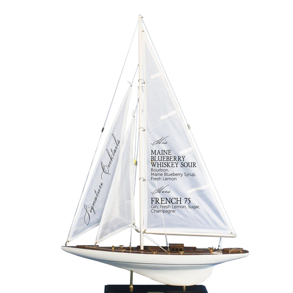A model of a sailboat with a white sail and brown deck, featuring text on the sail that reads "MAINE BLUEBERRY WHISKEY SOUR, Maine Blueberry Syrup, Fresh Lemon".