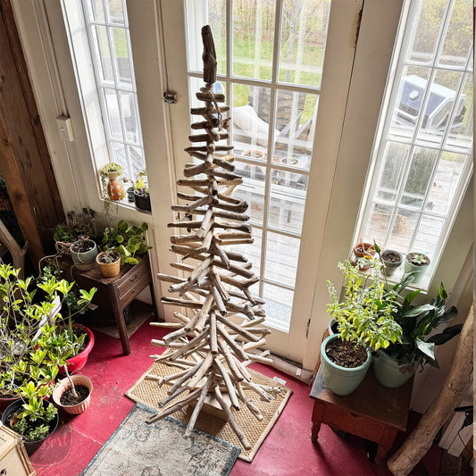 a rustic wooden Christmas tree made from driftwood, placed in a room with large windows overlooking a garden. The tree is surrounded by various potted plants, creating a cozy and natural atmosphere.