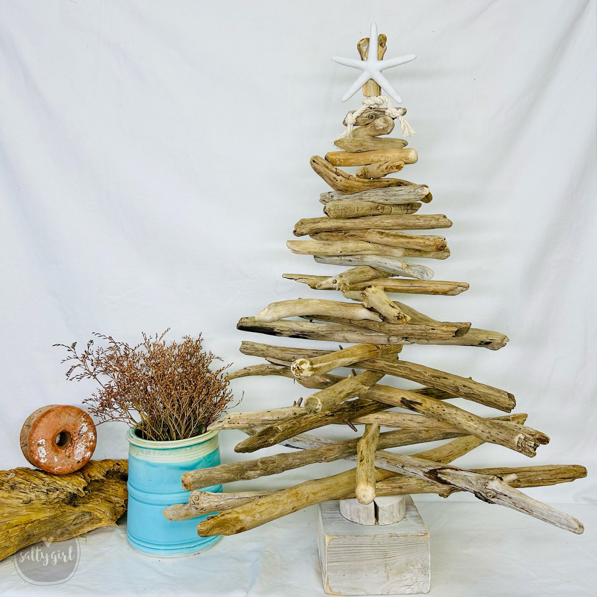 A rustic driftwood Christmas tree made from various branches and a star, placed on a white surface. Next to the tree is a blue pot containing dried flowers, and a donut is also visible in the background.