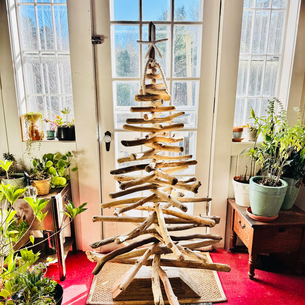A rustic driftwood Christmas tree stands in the center of a room, surrounded by potted plants and a large window.
