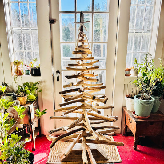 A rustic driftwood Christmas tree stands in the center of a room, surrounded by potted plants and a large window.