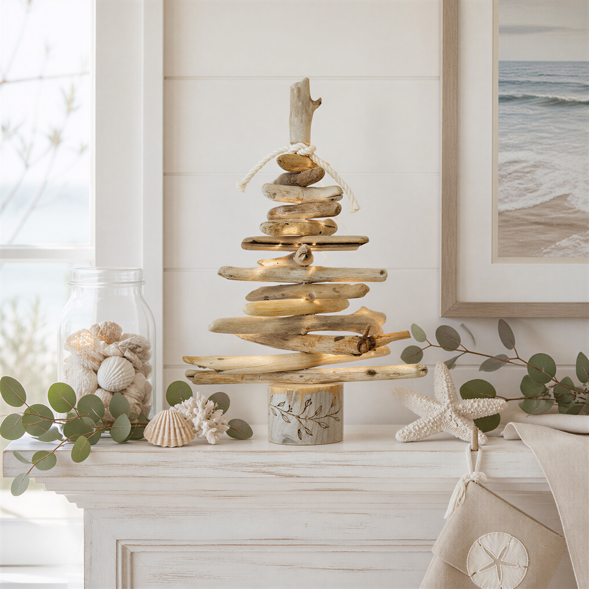 a rustic, driftwood Christmas tree made from stacked wooden planks, placed on a white wooden shelf. The tree is adorned with seashells and greenery, creating a coastal, beachy atmosphere.
