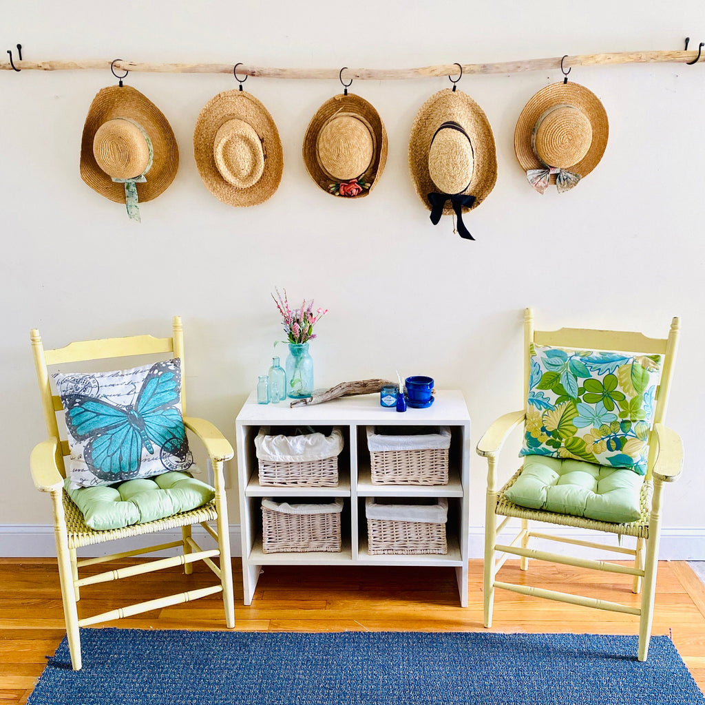 a cozy and inviting living space with a white wall, two yellow chairs, a white storage unit, and a blue rug. Above the chairs, there are five straw hats hanging on a wooden rack, adding a touch of rustic charm to the room. The storage unit has two shelves, providing ample storage space for the room's occupants. The arrangement of the furniture and accessories creates a harmonious and visually appealing space.