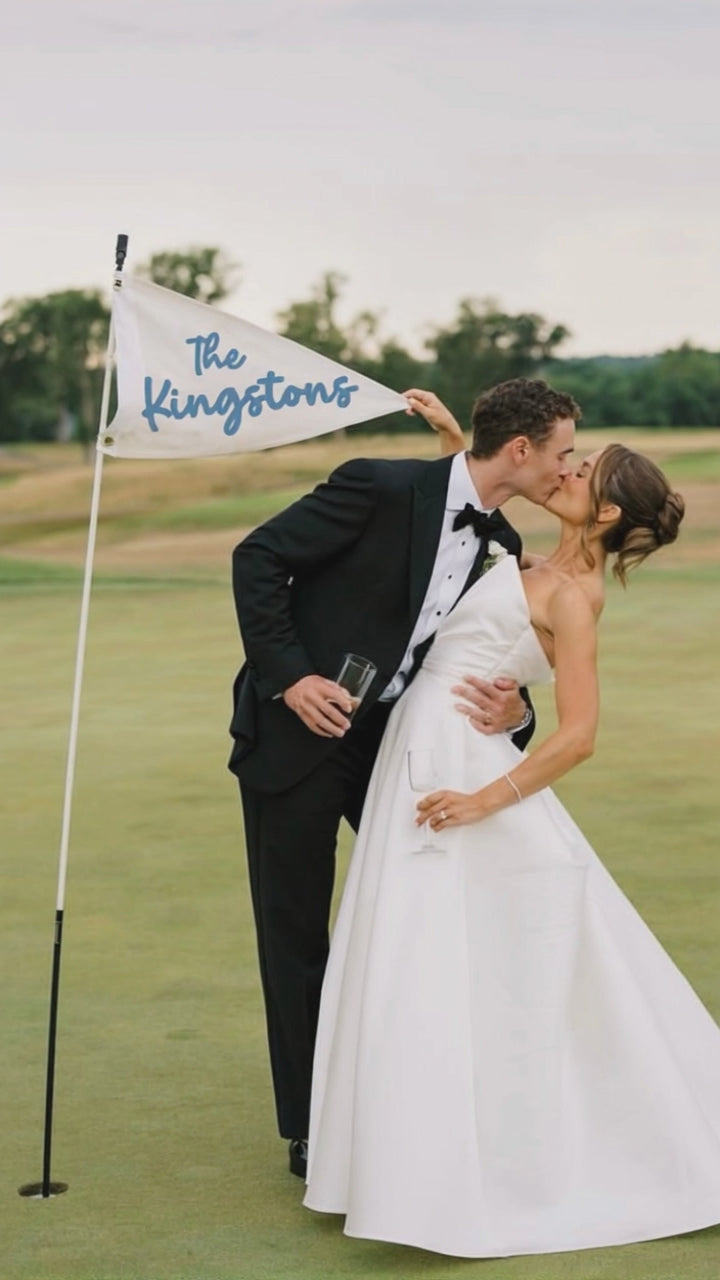 Couple in wedding attire kissing on a golf course with a flag reading 'The Kingstons'