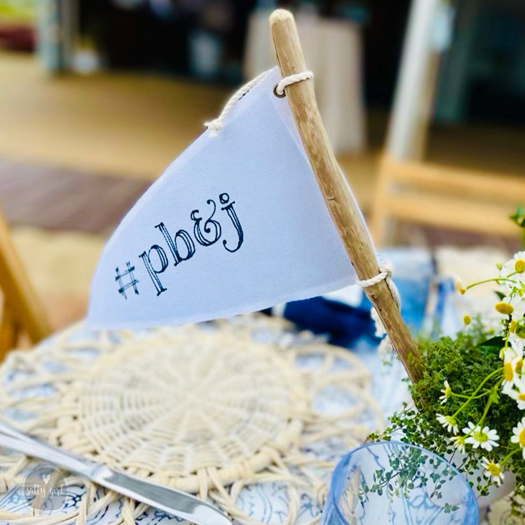 a table setting with a white tablecloth, a white plate, a blue vase with white flowers, and a small white flag with the text "#p&b&g" attached to it.