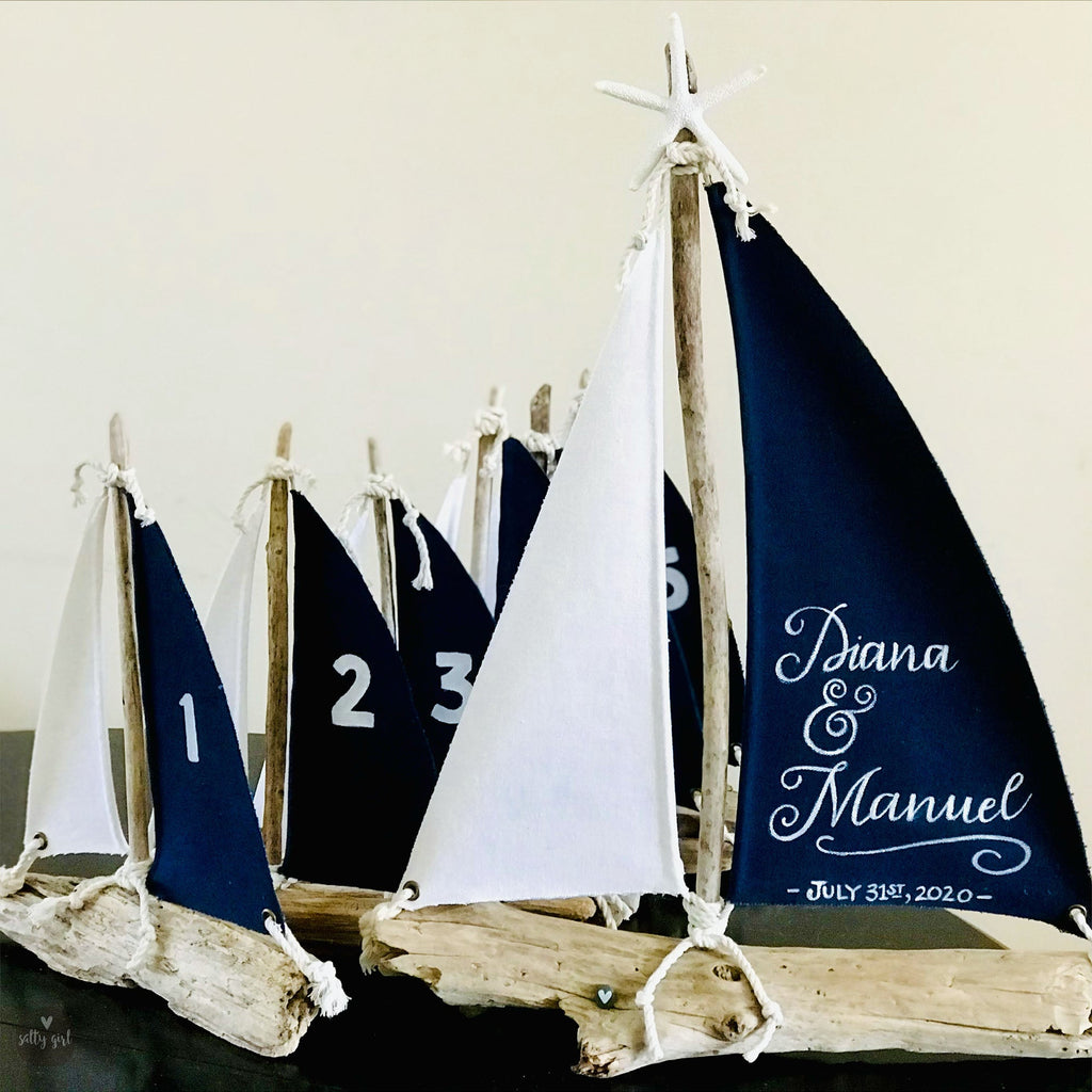 A collection of small sailboat-shaped decorations, including a large sailboat with the names Diana and Manuel written on it, displayed on a wooden base.