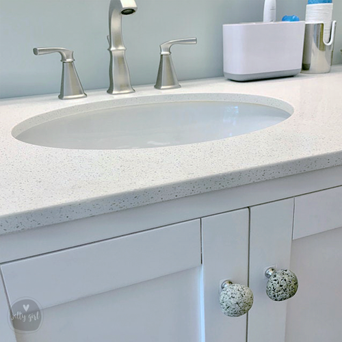 a bathroom sink with a white countertop and a silver faucet. The sink is set into a white cabinet with two round knobs. The background features a light blue wall with a white towel rack.