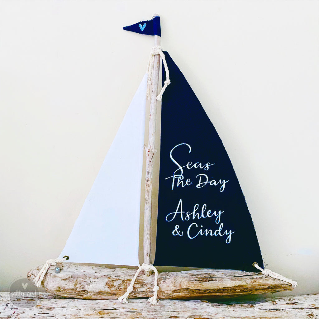 A handcrafted driftwood sailboat with a black and white sail, displaying custom lettering, a blue fabric flag on top, and affixed to a piece of driftwood.