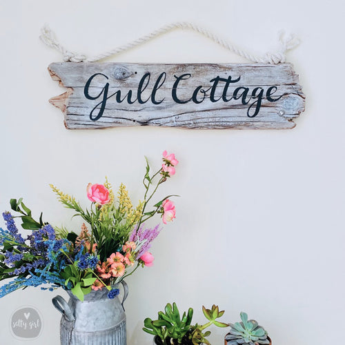 a rustic wooden sign that reads "Gull Cottage" hanging on a white wall, with a vase of colorful flowers and potted plants placed below it.