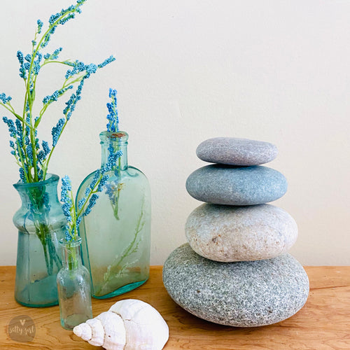 a wooden surface with three glass bottles, two blue and one green, and two blue and white vases with blue flowers. There are also three rocks, with the largest one being a gray stone and the other two being gray and white. A seashell is also present on the surface.