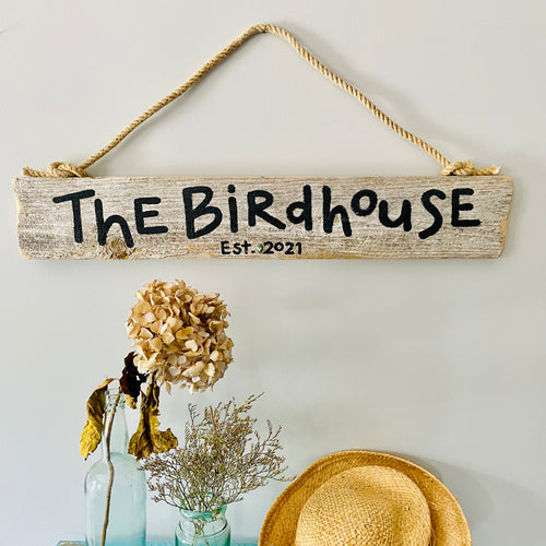 a rustic wooden sign that reads "The Birdhouse" hanging on a wall, accompanied by a dried flower arrangement and a straw hat.
